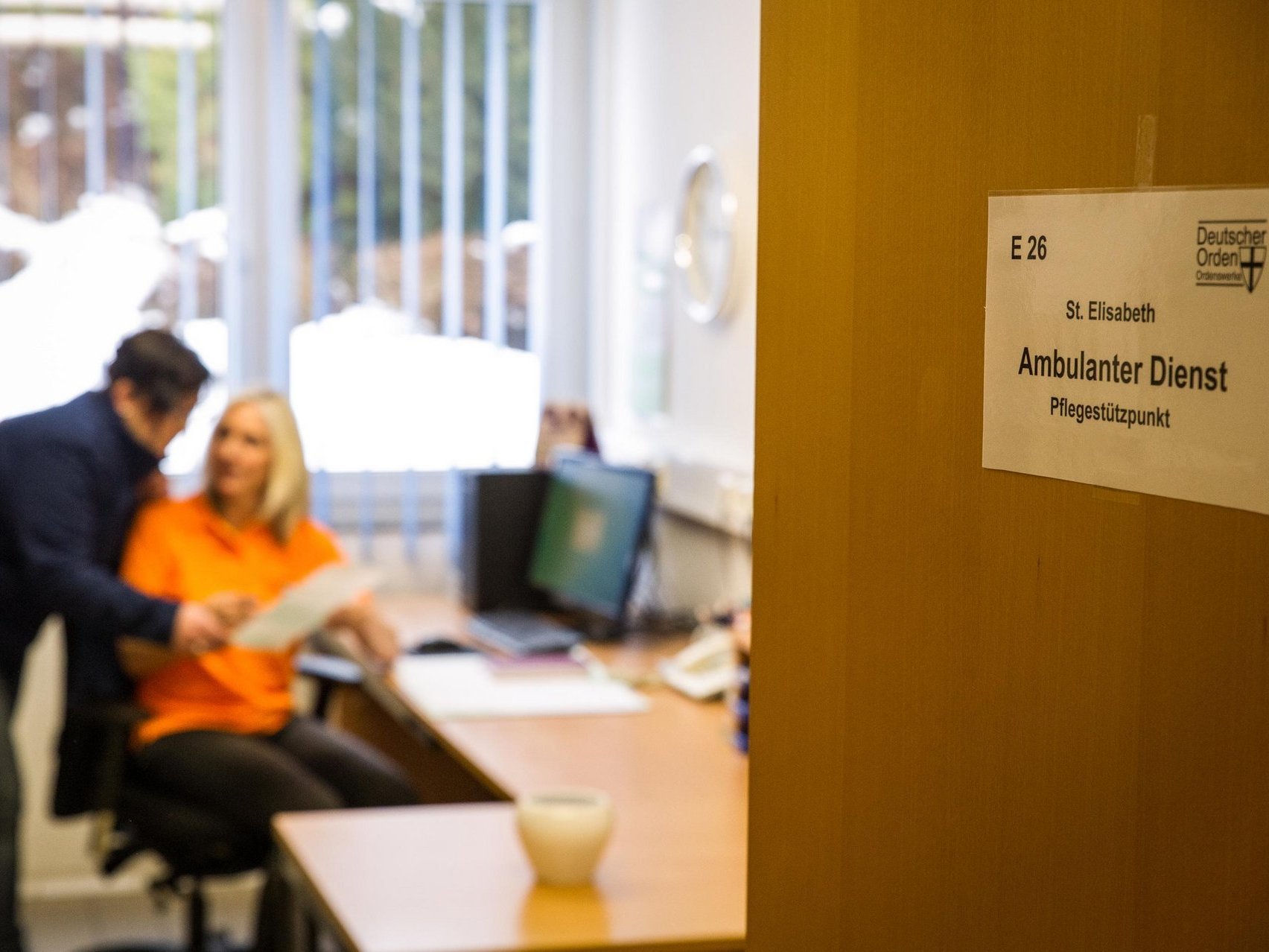 Auf einer halb geöffneten Bürotür sind auf einem Schild die Worte Ambulanter Dienst zu lesen. Im Inneren des Büros unterhalten sich zwei Personen über ein Schriftstück gebeugt.