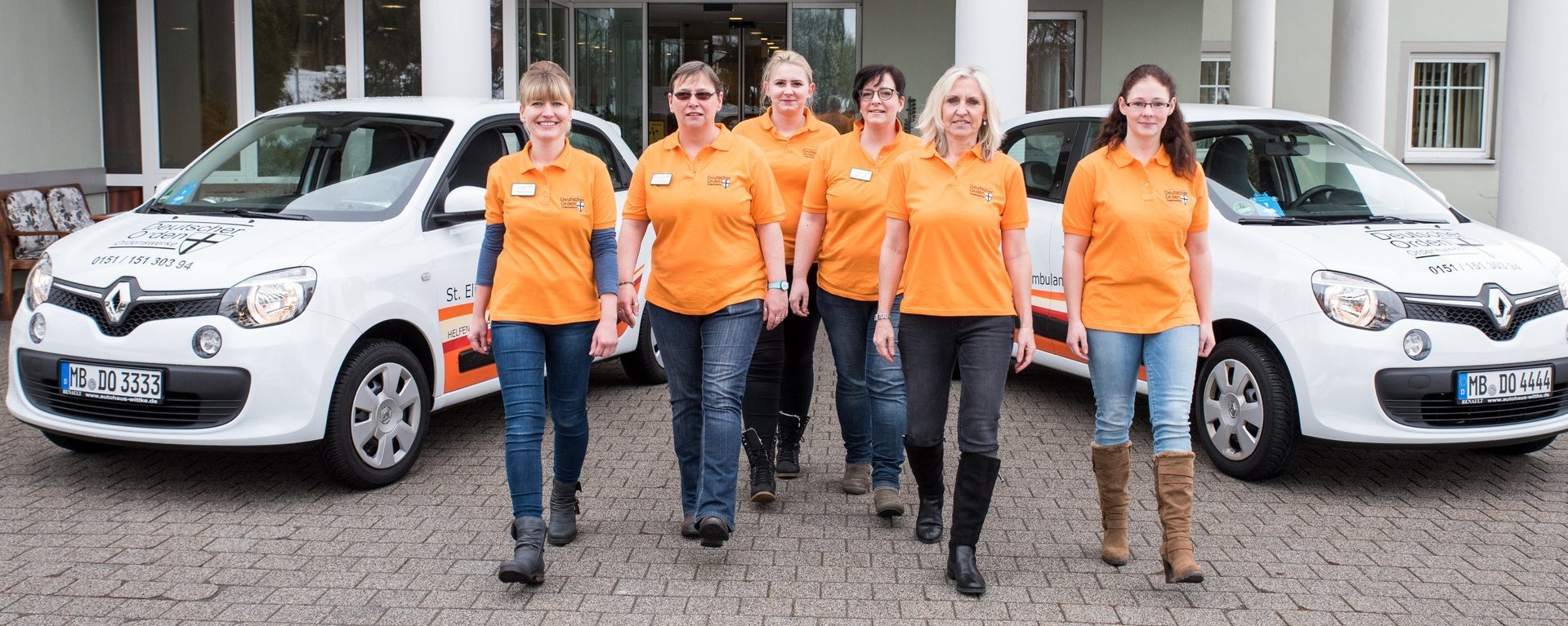 Vor einem Gebäudeeingang stehen zwei Fahrzeuge des Pflegedienstes Wunsiedel. Im Vordergrund gehen sechs Mitarbeiterinnen in orangenen T-Shirts lächelnd auf den Betrachter zu.