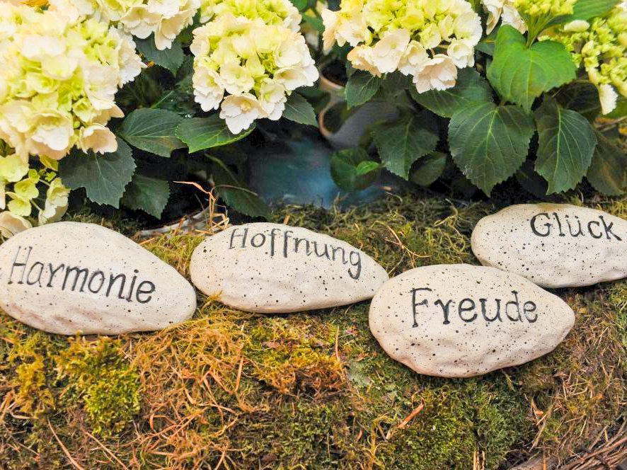 Vier ovale Steine liegen auf einem Moosbett vor einem Blumenbeet. Auf den Steinen stehen die Worte Harmonie, Hoffnung, Freude und Glück.